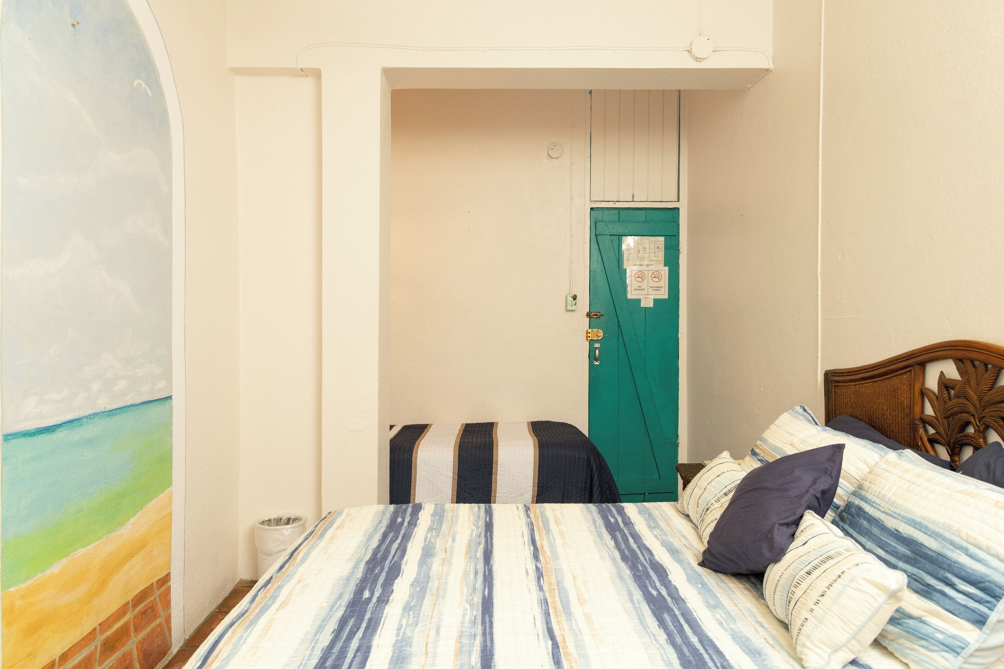 A bedroom with a bed, a green door, a wall with a painting, and a small trash bin.