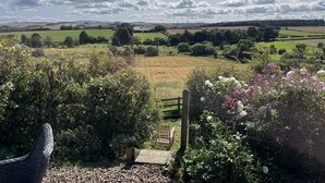 Un jardin avec un banc en bois, un chemin de gravier et une vue sur un paysage rural composé de champs et d’arbres.