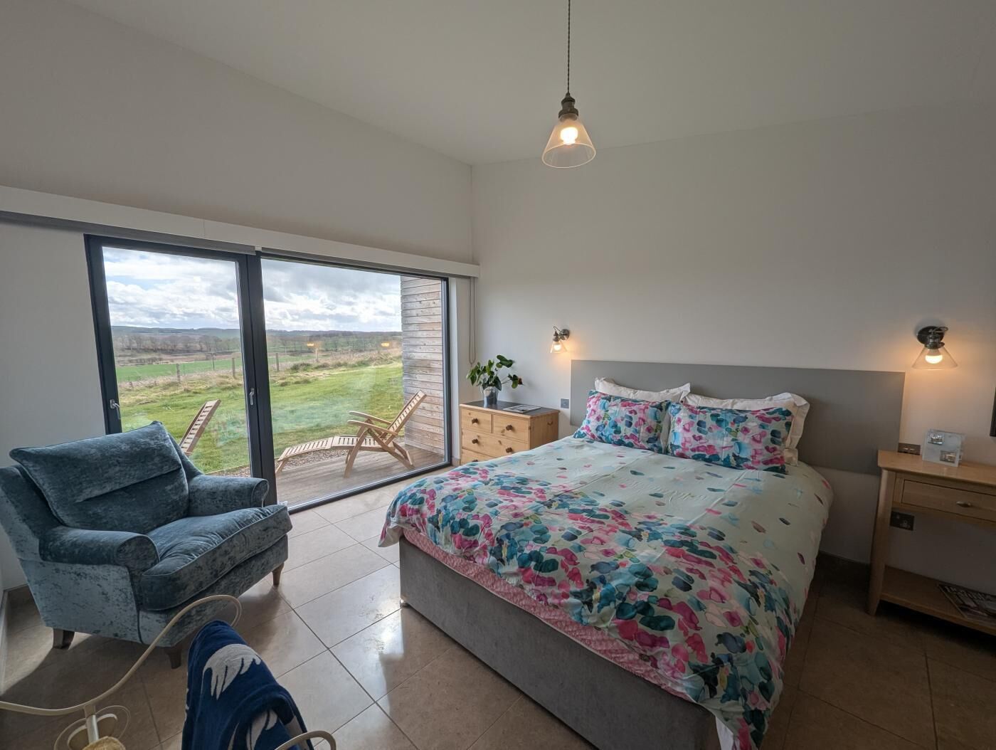 Un dormitorio con una cama grande, una silla, una mesita de noche y vistas al exterior a través de una puerta corredera.