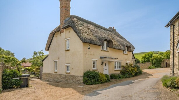 Ein traditionelles reetgedecktes Cottage mit einem Steinkamin, einer Schotterauffahrt und einer Steinmauer.