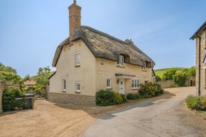 A traditional thatched-roof cottage with a stone chimney, a gravel driveway, and a stone wall. - 3 Bed in West Lulworth (oc-s32114) (West Lulworth)