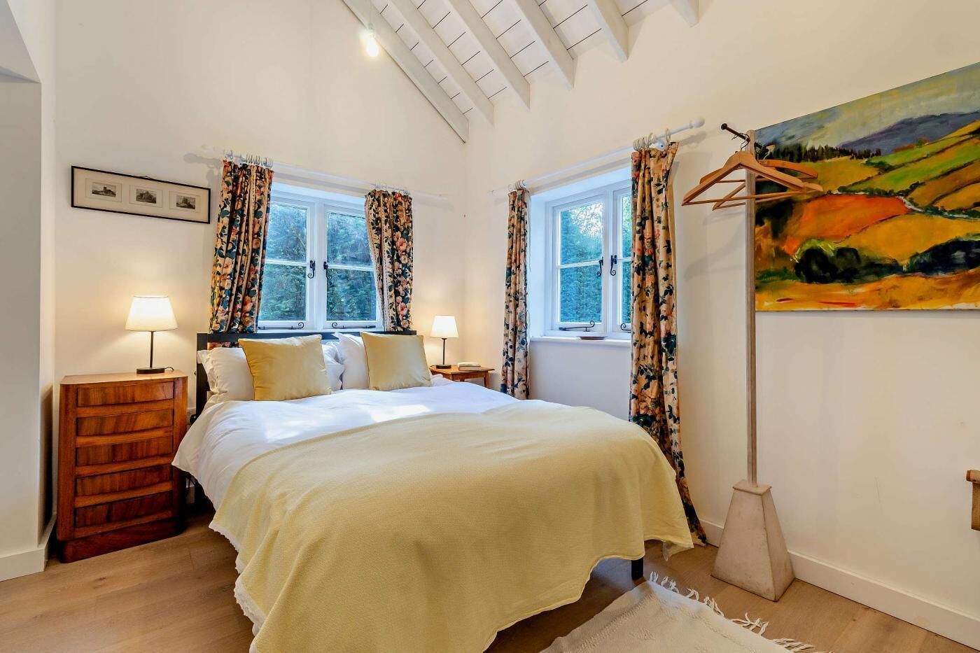 A bedroom with a bed, two bedside tables, a wooden dresser, a painting, and two windows with curtains.