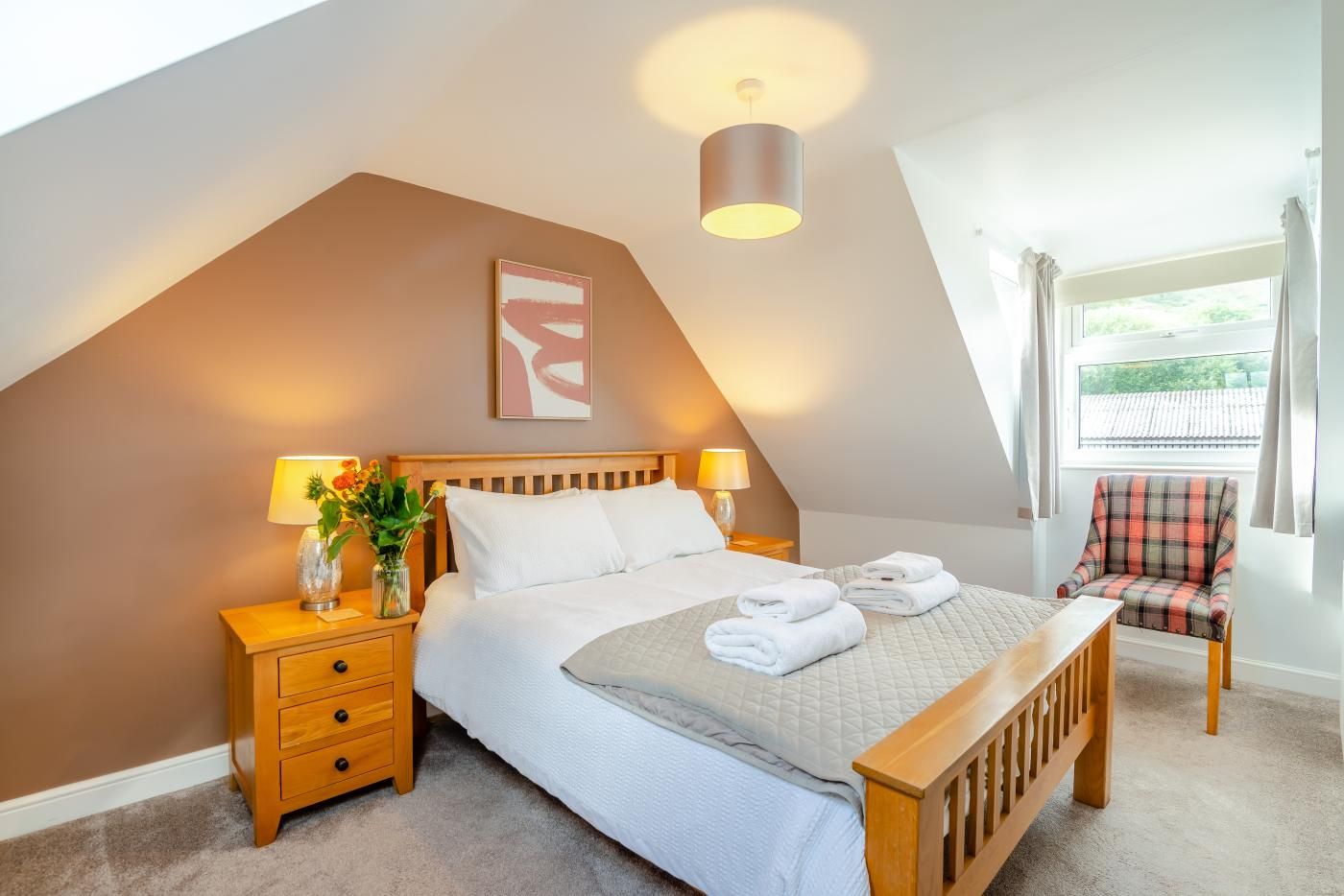 Een slaapkamer met een schuine kap, een bed met witte lakens, twee bedlampen, een stoel en een raam met gordijnen.