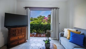 Ein Wohnzimmer mit Fernseher auf einer Holzkommode, einer Couch mit Kissen, einem Couchtisch mit Pflanze und Blick auf einen Garten durch eine Schiebetür.