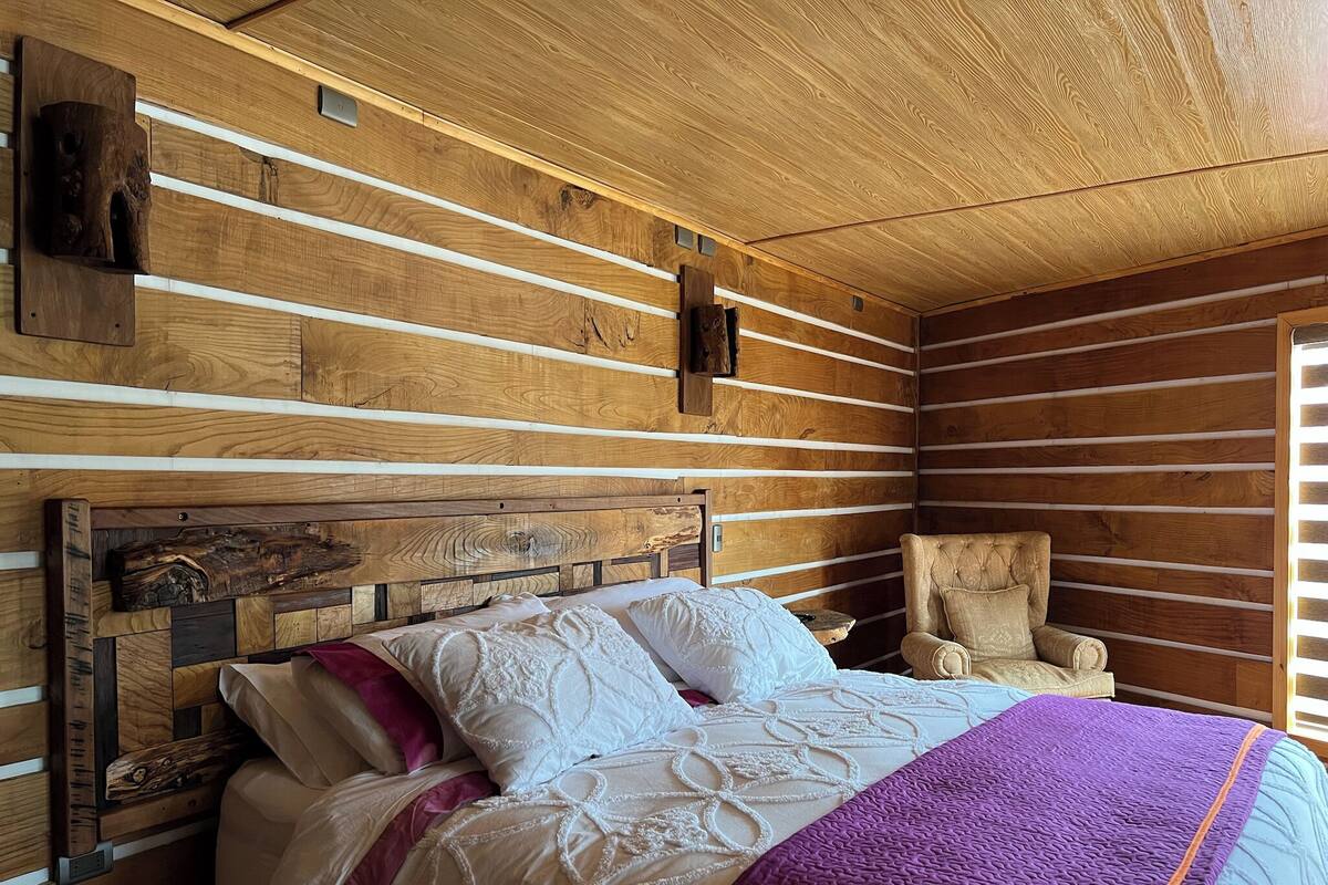 Een houten blokhutinterieur met een bed, kussens, een stoel en een houten hoofdbord.