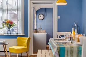 A room with a blue wall, a yellow chair, a table with a blue tablecloth, and a clock on the wall. - 3 Bed in Cromer (oc-2077) (Cromer)