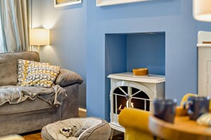 A cozy living room with a grey sofa, patterned cushions, a white fireplace, and a pet bed. - 3 Bed in Cromer (oc-2077) (Cromer)
