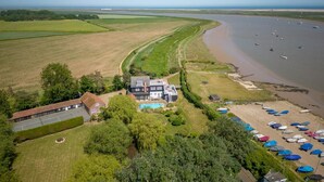 Vue aérienne d’une propriété située sur la côte, avec une grande maison, une piscine et une plage parsemée de parasols.