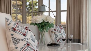 Un coin salon confortable avec un canapé, des coussins à motifs, un vase de fleurs, une carafe à vin et deux verres à vin posés sur une table.