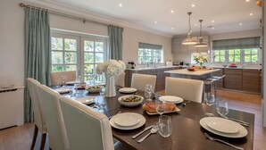 Un espace de repas moderne avec une grande table dressée pour un repas, une cuisine en arrière-plan et de grandes fenêtres laissant entrer la lumière naturelle.
