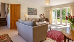 A cozy living room with a blue sofa, a brown armchair, and a red patterned rug. There is a wooden door, a painting on the wall, and a table with a vase of flowers. - 2 Bed in Kettlestone (oc-t32268) (Little Snoring)