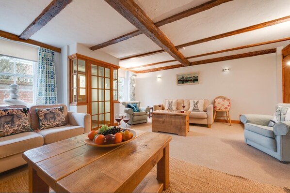 A cozy living room with a wooden coffee table, a sofa, and a chair. There is a window with curtains and a painting on the wall. - 4 Bed in Wells-next-the-Sea (oc-2045) (Wells-next-the-Sea)
