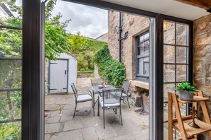 An open patio with a stone table and chairs, a potted plant, and a view of a stone building with a door and greenery. - 4 Bed in Alnwick (oc-g32272) (Alnwick)