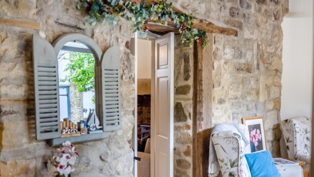 Une pièce aux murs de pierre, avec une chaise blanche, un miroir à volets et une porte donnant sur une autre pièce.