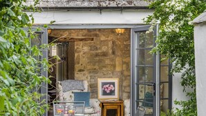 Un espace de détente extérieur accueillant, avec un mur en pierre en arrière-plan, un fauteuil à motifs floraux, une petite table avec des boissons et un tableau encadré.