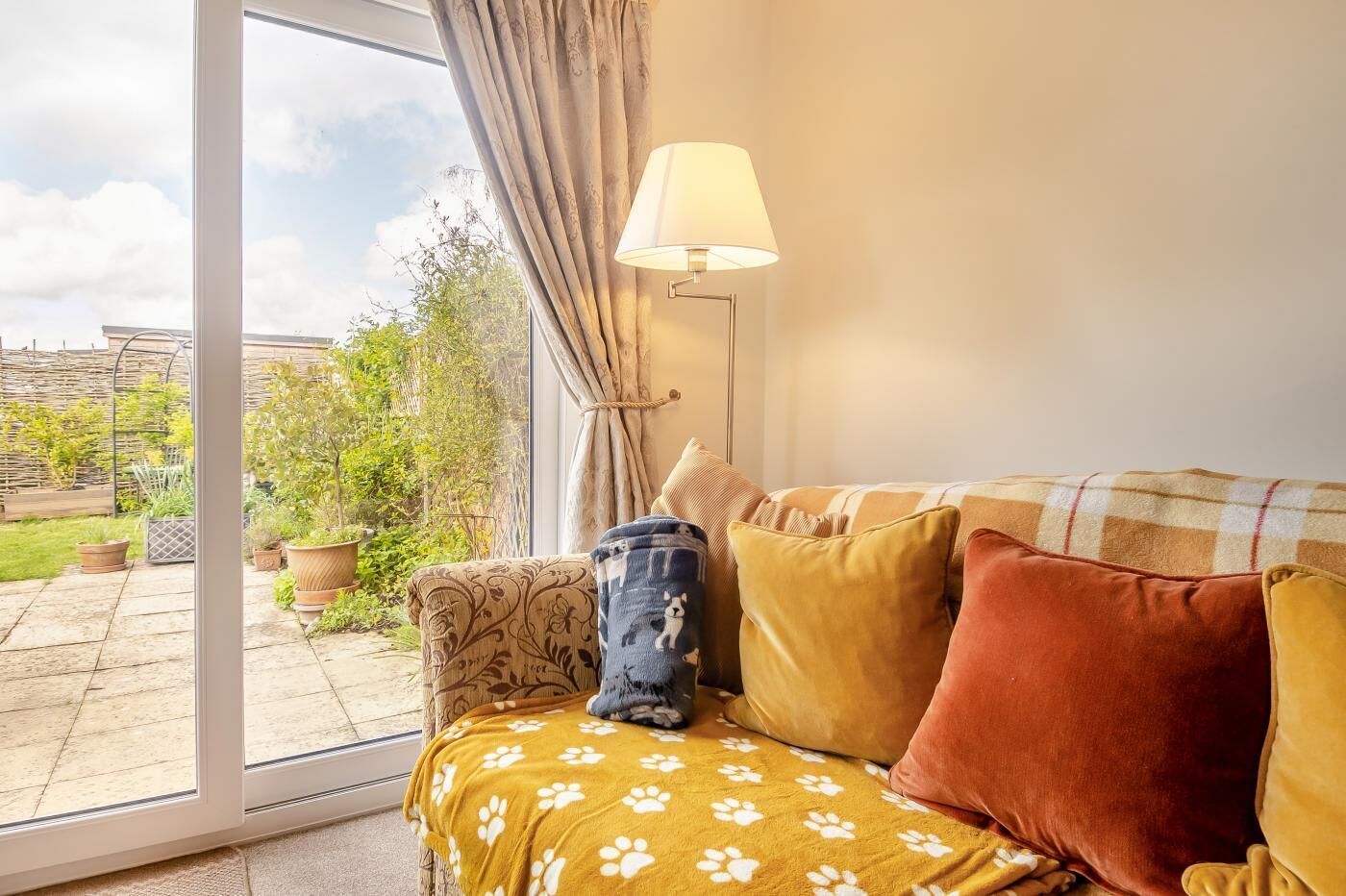 A cozy living room with a sofa, a lamp, and a view of a garden through a glass door.