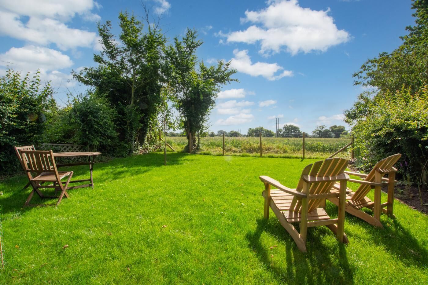 A grassy outdoor area with wooden chairs and a table, surrounded by trees and a fence.