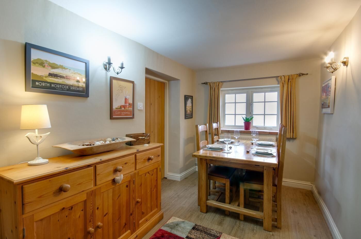 Un coin repas avec une table en bois dressée pour quatre personnes, un buffet en bois avec des tiroirs, une lampe et des tableaux encadrés accrochés au mur.