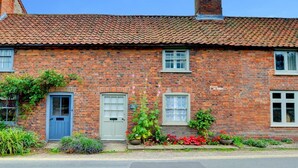 Une petite maison en briques avec un toit en tuiles, deux portes et un petit jardin agrémenté de fleurs et de plantes.