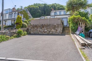 A paved driveway leading to a stone wall with a wooden gate, surrounded by greenery and residential buildings. - 4 Bed in Mumbles (oc-t32236) (The Mumbles)