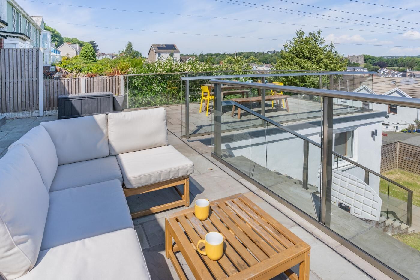 En balkon med en sofa, et træbord med to gule kopper og et gelænder af glas med udsigt over et boligområde.