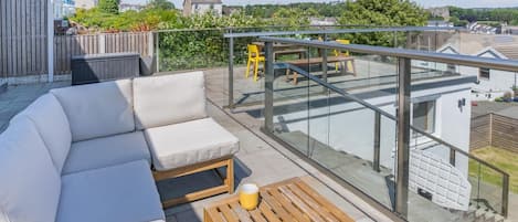A balcony with a sofa, a wooden table with two yellow cups, and a glass railing overlooking a residential area.