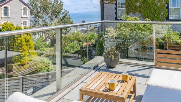 Ein Balkon mit Glastrennwand, ein Holztisch mit zwei gelben Tassen, ein Topfpflanze und weiß gepolsterte Sitzgelegenheiten.