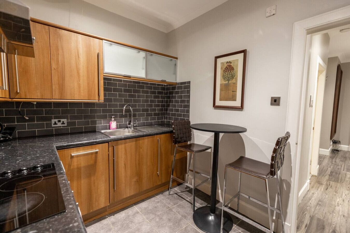 Een moderne keuken met houten kastjes, een zwart betegelde spatwand, een roestvrijstalen spoelbak en een kleine ronde tafel met twee stoelen.