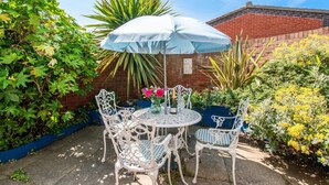 A patio with a round table and white chairs, an umbrella, and a vase with flowers. - 2 Bed in Sheringham (oc-2029) (Sheringham)