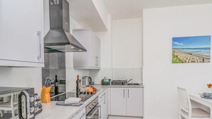 Een kleine keuken met witte kastjes, een gasfornuis, een magnetron en een eethoekje met een stoel en tafel.