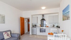Een knus woongedeelte met een kitchenette, een grijze bank, een witte eettafel en een witte stoel.