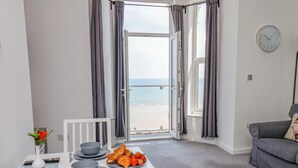 Een kamer met een ontbijt gedekte eettafel, een grijze bank en uitzicht op het strand door open gordijnen.