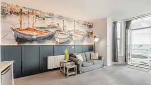 Uma sala de estar moderna com um quadro grande de barcos, um sofá cinzento, uma mesa pequena e vista para o mar através de uma janela ampla.