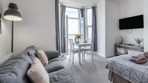 Ein modernes Hotelzimmer mit grauem Sofa, einem Esstisch mit Stühlen, einem Bett mit gemustertem Überwurf, einem an der Wand montierten Fernseher und einem großen Fenster mit Vorhängen.