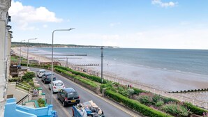 Eine Küstenstraße mit parkenden Autos, einer Promenade und Blick auf Strand und Meer.
