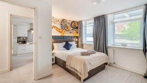 Une chambre à coucher avec un lit, une tête de lit, une fenêtre avec des rideaux et une cuisine dans la pièce adjacente.