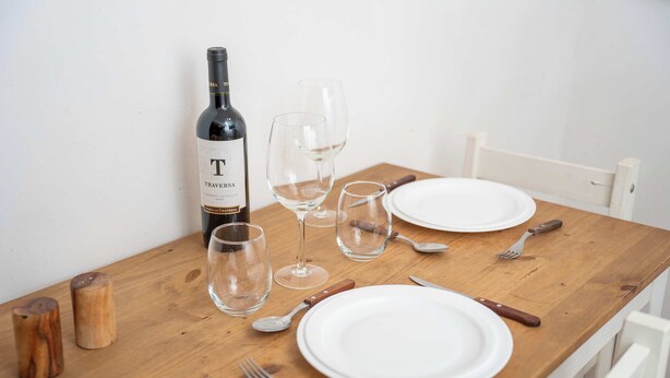Una mesa de madera dispuesta para comer, con una botella de vino, copas, platos, cubiertos y saleros y pimenteros.