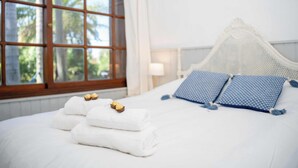A neatly made bed with white linens and two pillows, a window with curtains, and a view of greenery outside.