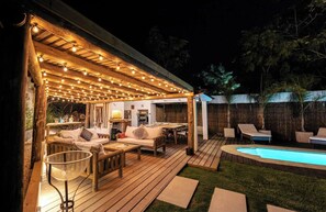 Un terrazzo in legno con mobili da esterno, una piscina e una zona relax coperta con luci a stringa.