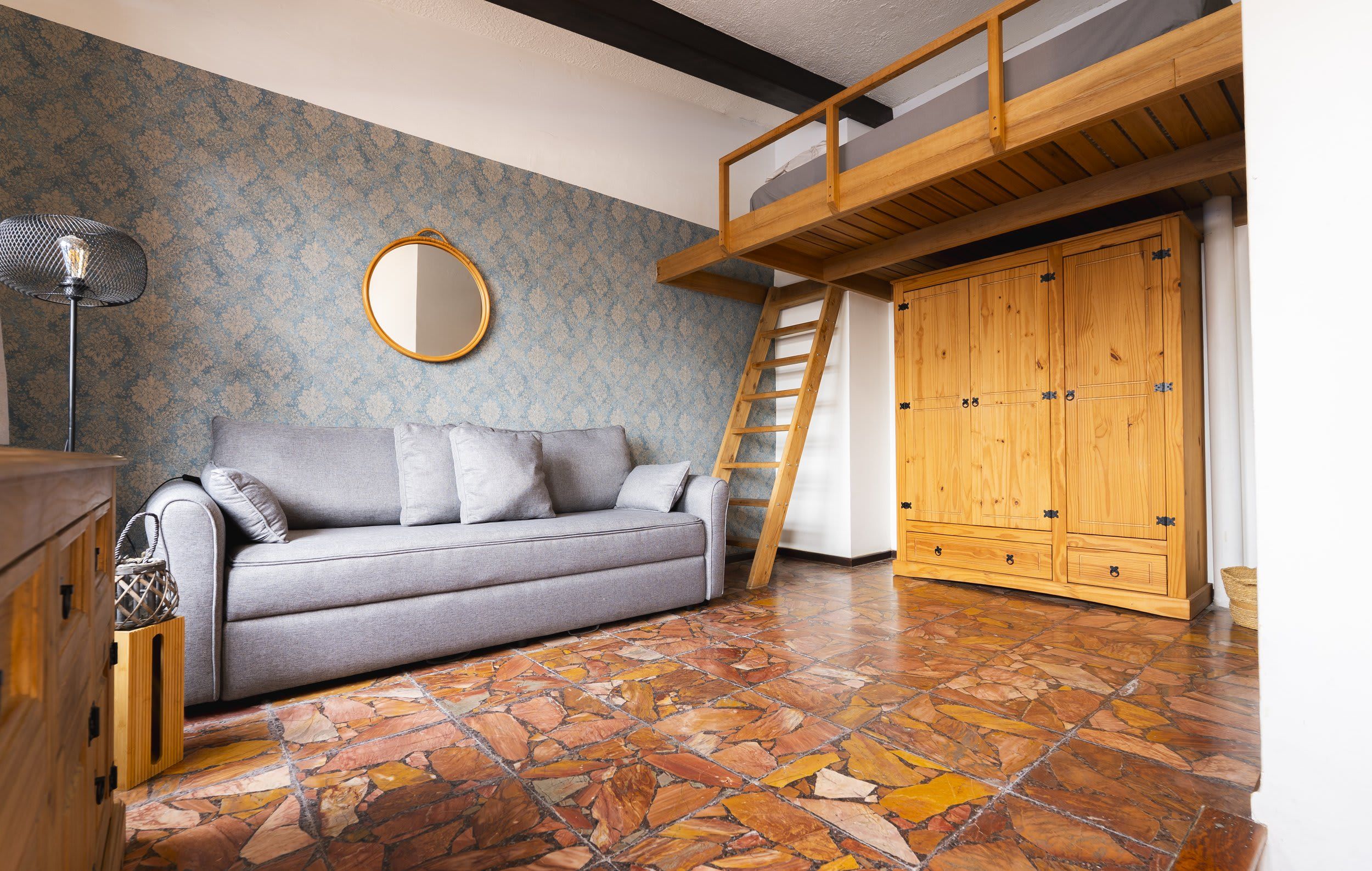 A room with a grey sofa, a wooden wardrobe, a mirror, and a stone-tiled floor.