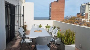 Eine Terrasse mit einem weißen Tisch und Stühlen, Blumentöpfen und Blick auf das Meer und die Bebauung.