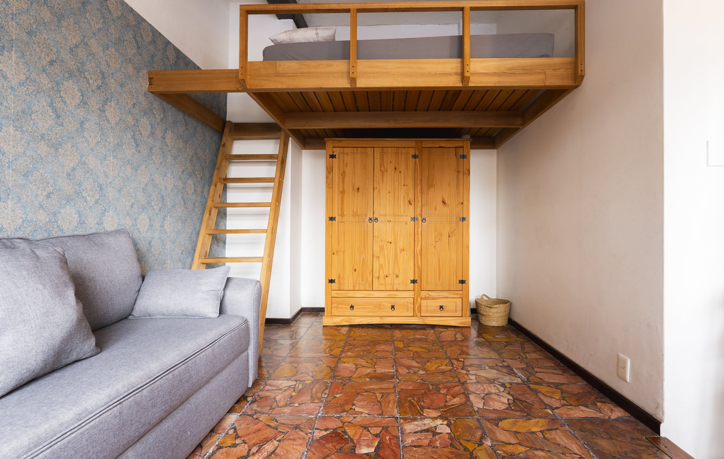 A loft bedroom with a bunk bed, a wooden wardrobe, a grey sofa, and a stone-tiled floor.