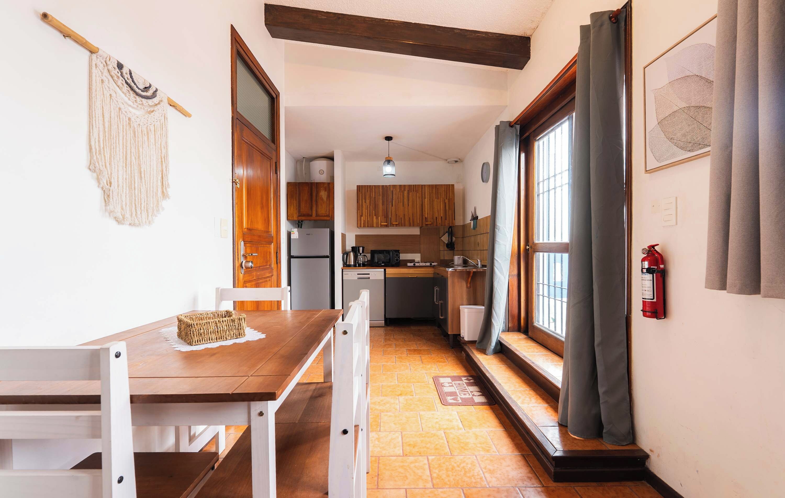 A narrow kitchen with wooden cabinets, a dining table with chairs, and a window with curtains.