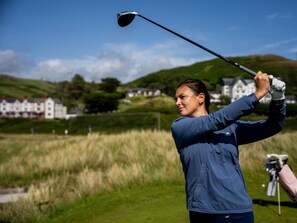 Une personne vêtue d’une veste bleue, en train de faire un swing avec un club de golf sur un terrain de golf, avec des maisons en arrière-plan.