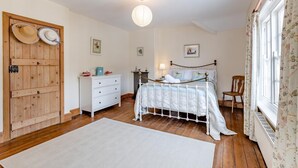 Ein Schlafzimmer mit Holztür, weißem Schminktisch, einem Bett mit Metallgestell und einem Fenster mit floralen Vorhängen.