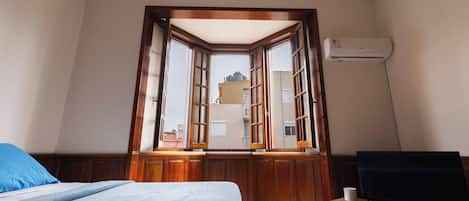 A bedroom with a bed, a television, and a window with a view of buildings.
