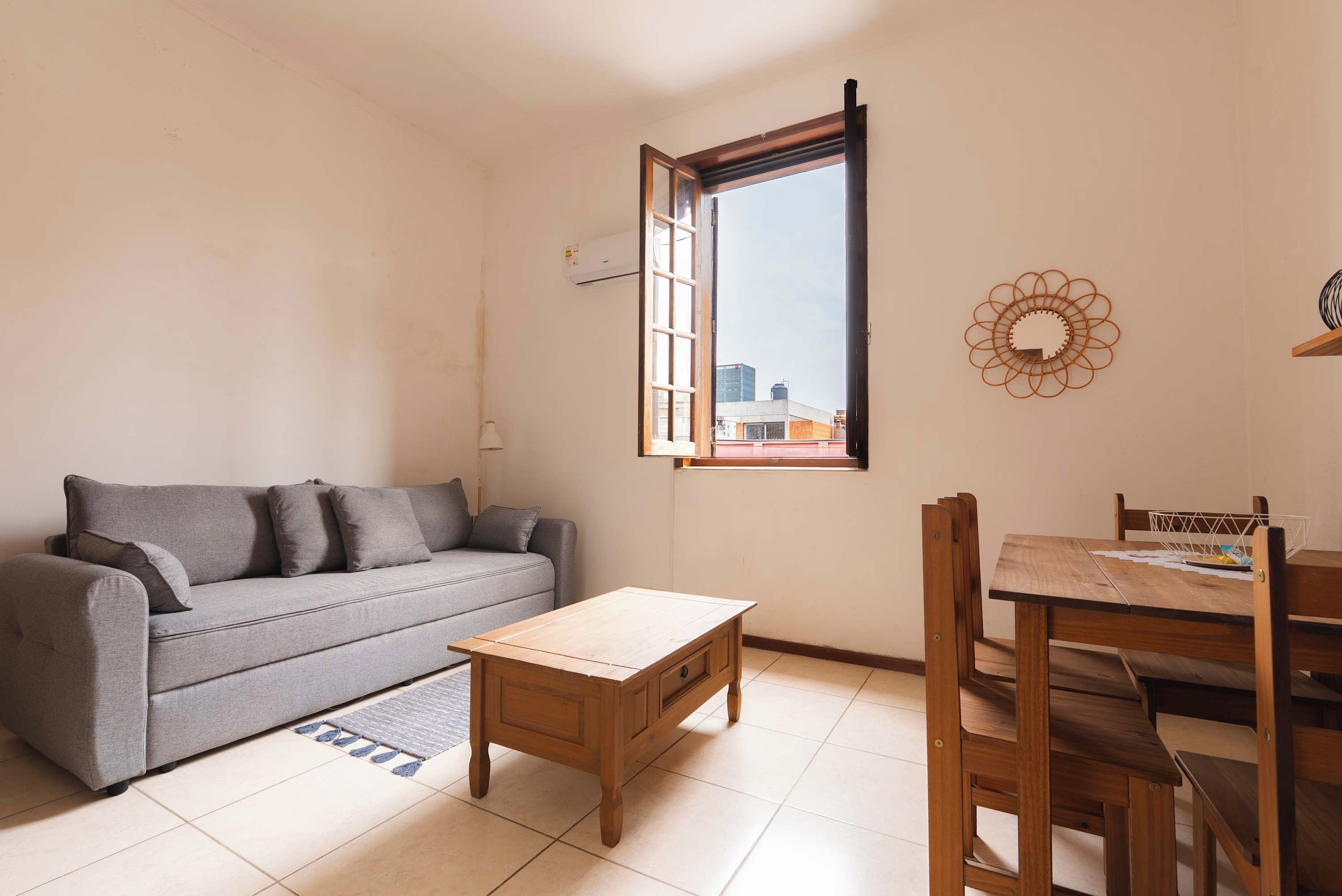 A living room with a grey sofa, a wooden coffee table, a dining table with chairs, and a window with a view of buildings.