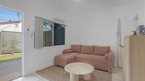 A room with a beige sectional sofa, a round wooden coffee table, a white air conditioning unit, a window with blinds, and a door leading outside. - Unit tucked away in a quiet street (Amity)