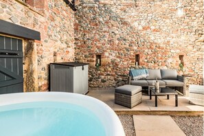 Un espace extérieur entouré de murs en pierre, avec un jacuzzi, un coffre de rangement gris, un canapé gris et une petite table recouverte d’une couverture.