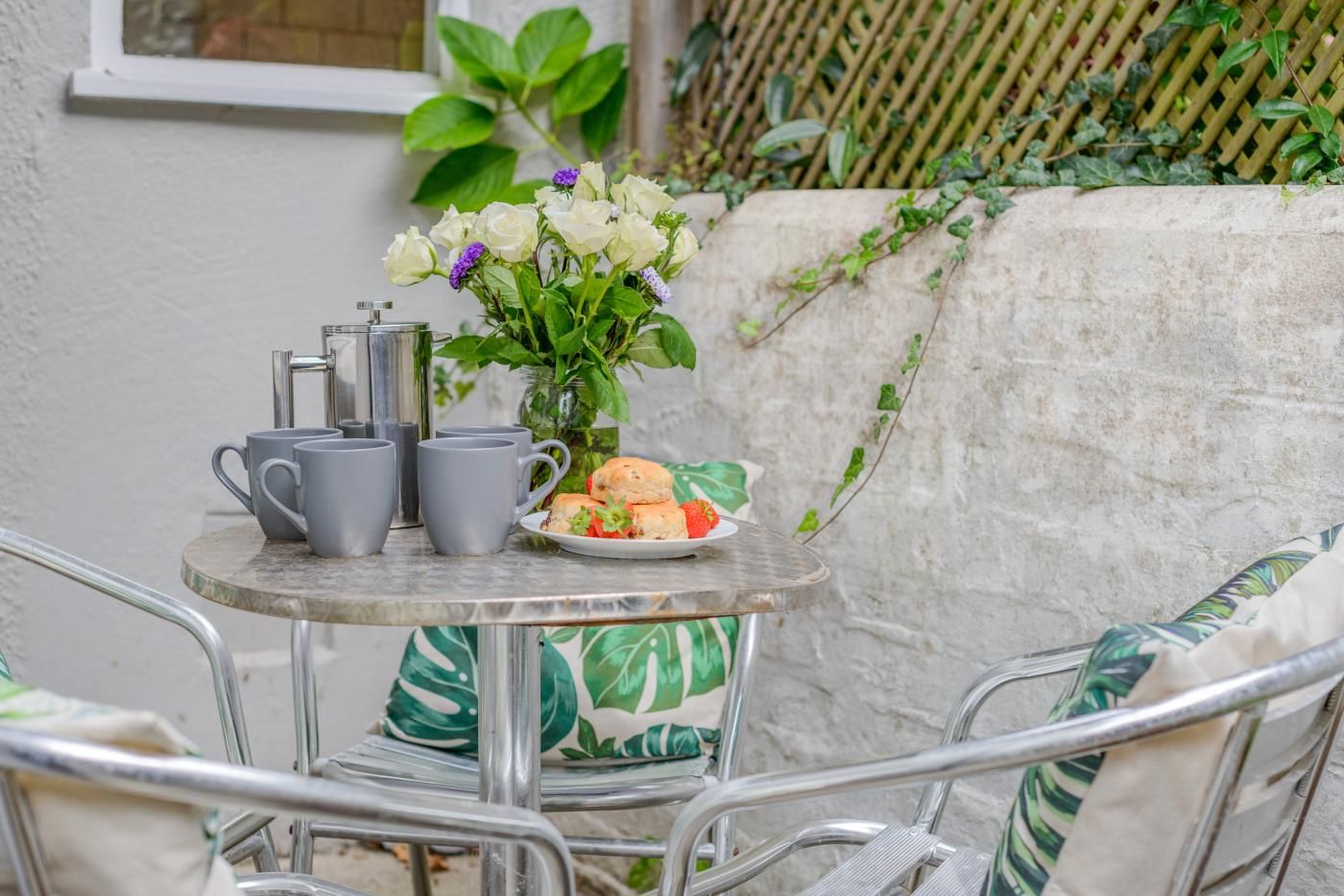 A metal table with grey cups, a glass pitcher, and a plate with food, set against a white wall with greenery.